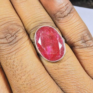 May include: A silver ring featuring a large, oval-shaped ruby gemstone. The ruby is a deep red colour and has a slightly textured surface.