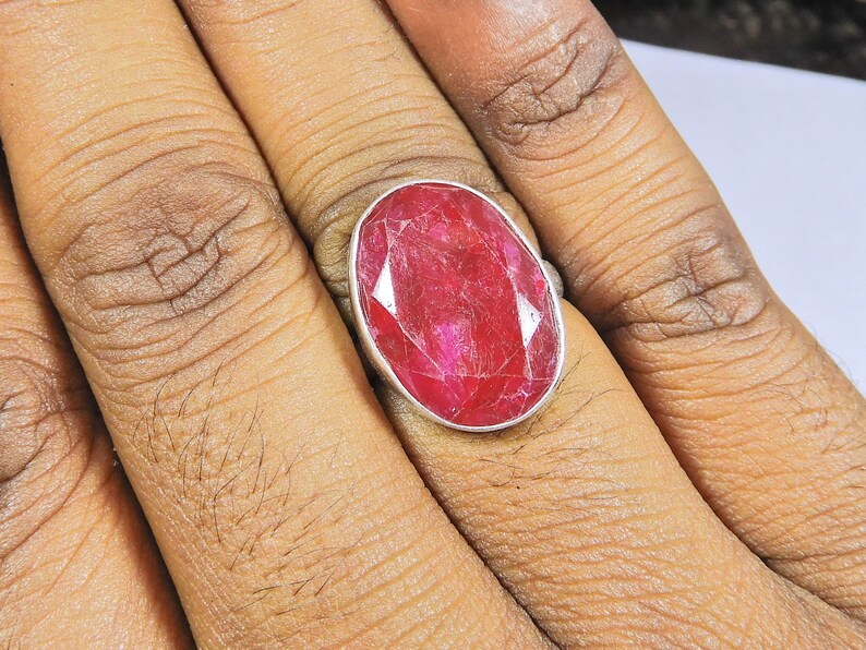 May include: A silver ring featuring a large, oval-shaped ruby gemstone. The ruby is a deep red colour and has a slightly textured surface.