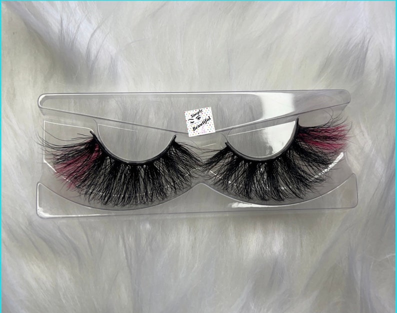 May include: A pair of false eyelashes in a clear plastic case. The eyelashes are black with a pink accent on the outer corner. The case has the text "Simply Beautiful" printed on it.