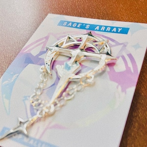Può includere: Uno spillo in metallo argentato con un design a forma di luna crescente con una stella e una catena. Lo spillo è su una carta bianca con il testo "Sage's Array".