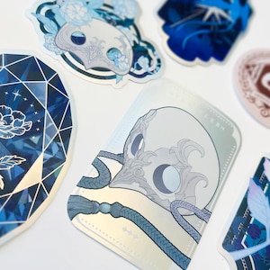 Könnte beinhalten: Eine Sammlung dekorativer Aufkleber mit floralen und geometrischen Designs in Blau-, Weiß- und Silbertönen. Die Aufkleber zeigen detaillierte Illustrationen von Blumen, Schädeln und abstrakten Mustern mit glänzendem Finish.