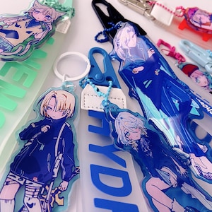 May include: Collection of anime-style acrylic keychains featuring colorful illustrations of characters. The keychains have various designs, including characters with cat ears and long hair. Some keychains have text in English and Japanese.
