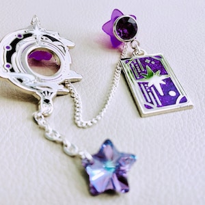 May include: A silver charm bracelet with a purple star charm, a silver charm with a purple and white design, and a silver chain. The charm with the purple and white design has a star in the centre.