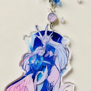 Könnte beinhalten: Ein blauer und weißer Acryl-Schlüsselanhänger mit einem sich umarmenden Paar. Die Frau trägt ein rosa Kleid und der Mann ein blaues und schwarzes Outfit. Der Schlüsselanhänger hat eine silberne Kette und einen glitzernden Anhänger.