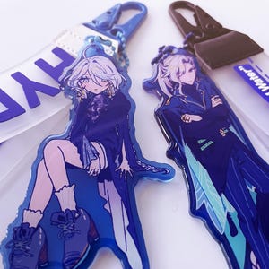 Op de afbeelding: Twee acryl sleutelhangers in anime-stijl met personages in blauwtinten. Eén sleutelhanger heeft het woord "HYD" op de band, de andere "Archon of Water". Beide hebben blauwe hardware.