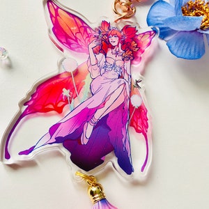 May include: A pink and purple fairy with red hair and wings sits on a flower. The fairy is wearing a white dress and has a gold crown. The fairy is part of a charm with a pink tassel.