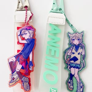 May include: Two keychains with anime-style illustrations. One keychain features a character in a red and blue outfit, the other has the word "ANEMO" in green. Both have a white strap and a metal clasp.