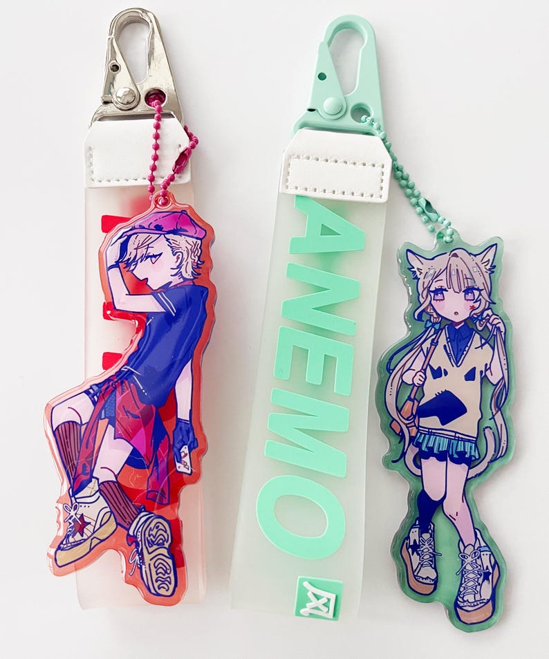 May include: Two keychains with anime-style illustrations. One keychain features a character in a red and blue outfit, the other has the word "ANEMO" in green. Both have a white strap and a metal clasp.