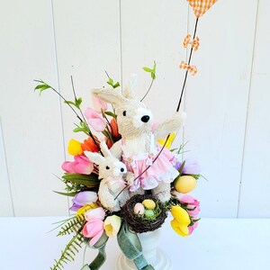 May include: A white ceramic vase with a green ribbon, filled with a spring floral arrangement featuring pink, yellow, and purple tulips, green ferns, and two bunny figurines. One bunny is holding a kite with a gingham pattern.