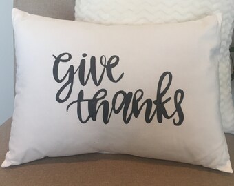 May include: White throw pillow with black cursive text that reads "give thanks" on a beige chair.