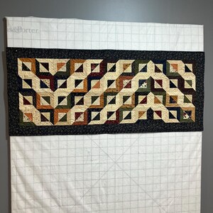 May include: A quilt with a geometric pattern in shades of brown, green, cream, and black. The quilt is hanging on a wall with a gridded background.