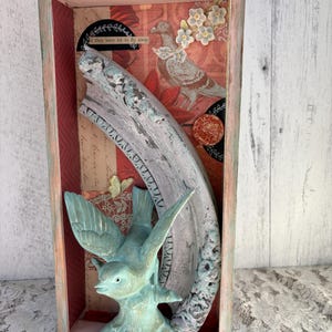 May include: A rectangular shadow box with a distressed, painted frame. Inside, a vintage-style collage features a teal bird sculpture, a curved, ornate frame piece, and a printed background with a pigeon and floral accents. The text "if they were let to fly away" is visible.