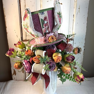 May include: A decorative Easter-themed arrangement featuring a top hat with faux bunny ears, adorned with floral patterns, pearls, and ribbons. The arrangement includes colorful faux flowers, burlap ribbon, and a white vase, creating a festive display.