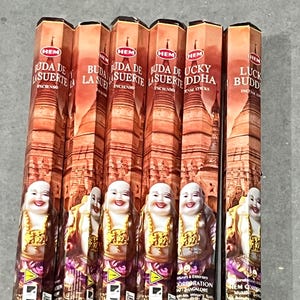 May include: Six boxes of incense sticks with a red, white, and gold design. The boxes feature an image of a smiling Buddha and the text "Buda de la Suerte" and "Lucky Buddha".