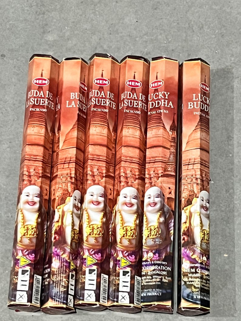 May include: Six boxes of incense sticks with a red, white, and gold design. The boxes feature an image of a smiling Buddha and the text "Buda de la Suerte" and "Lucky Buddha".