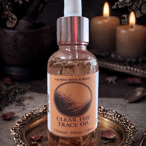 May include: A clear glass bottle with a white dropper top, labeled "SACRED ROSE & ROOT" and "CLEAR THE TRACE OIL." The label features a circular design. The bottle sits on a decorative gold tray with dried rose petals. Two lit candles are in the background.