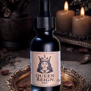 May include: A black glass bottle of "Queen Reign Oil" with a label featuring a crowned woman. The bottle has a dropper and is set on a gold-coloured tray with dried rose petals. The text "Sacred Rose & Root" is at the top.