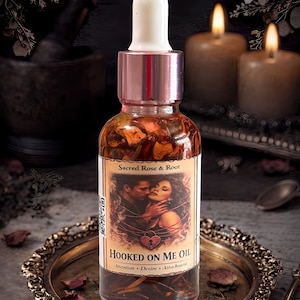 May include: A clear glass bottle of "Hooked On Me Oil" with a white dropper top and a rose gold cap. The label reads "Sacred Rose & Root" and features an image of a couple. The bottle sits on a decorative tray with dried rose petals.