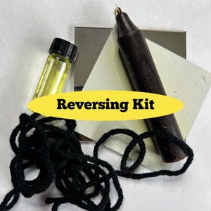 Pode incluir: Uma vela preta, um pequeno frasco de líquido amarelo, um cordão preto e um pequeno espelho prateado. Os itens estão dispostos sobre uma superfície branca. O texto "Reversing Kit" está escrito em amarelo sobre uma faixa preta.