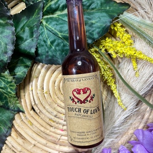 Puede incluir: Una botella de vidrio marrón con una tapa negra y una etiqueta que dice "Touch of Love Patchouli" de Murray & Lanman. La botella está sentada en una cesta tejida con flores moradas en primer plano.