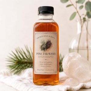 Può includere: Una bottiglia di vetro trasparente con tappo nero, riempita di liquido ambrato. L'etichetta recita "Sacred Rose & Root Pine Tar Wash" con un'illustrazione di pigna. Su un panno bianco, con un cristallo di quarzo.