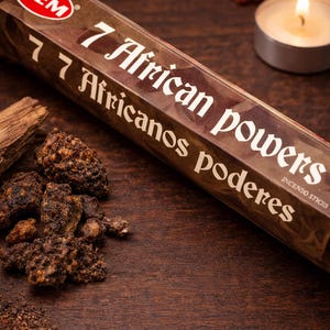 May include: A brown incense stick box with the text "7 African Powers" and "7 Africanos Poderes" is displayed on a wooden surface. Beside the box are incense sticks, a lit tea light candle, and other aromatic materials.