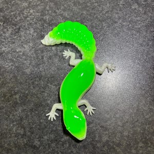 May include: A vibrant green toy lizard with a translucent, gel-like body. The lizard features small, white feet and a curved tail. The body is a bright green, and the tail is a lighter shade of green. The toy is on a dark grey surface.