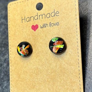 May include: A pair of black stud earrings with colorful, abstract designs. The earrings are displayed on a brown card with the text "Handmade with love" and a heart symbol.