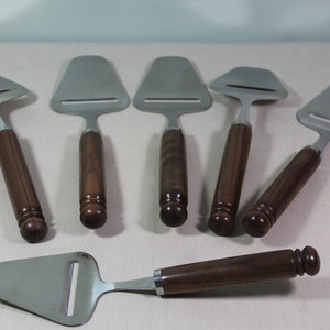 May include: Six stainless steel cheese slicers with brown wooden handles. The slicers are arranged in a row on a white surface.