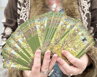Op de afbeelding: Een waaier van groene en bruine tarotkaarten met illustraties van bomen en vogels. De kaarten worden in de handen van een persoon uitgespreid.