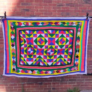 May include: A colourful patchwork quilt with a geometric pattern in shades of red, yellow, blue, green, and white. The quilt is hanging on a brick wall.