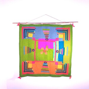 Op de afbeelding: Een kleurrijke wandhanger met een geometrisch patroon in tinten groen, blauw, oranje, roze en geel. De hanger is gemaakt van stof en heeft een houten lat aan de bovenkant.