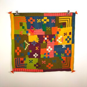 May include: A colourful patchwork quilt with a geometric design. The quilt is made up of squares of different colours and patterns, including green, orange, red, blue, and yellow. The quilt is hanging on a white wall.