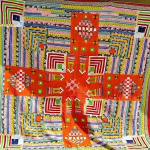 Puede incluir: Una colcha colorida con estampado geométrico con un esquema de colores rojo, naranja, amarillo, verde, azul y blanco. La colcha está colgada en un tendedero fuera de un edificio.