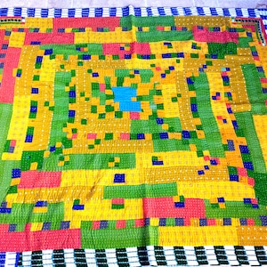 Op de afbeelding: Een kleurrijk patchwork-deken met een geometrisch ontwerp. Het deken is gemaakt van vierkanten in verschillende kleuren, waaronder geel, groen, rood en blauw. De vierkanten zijn gerangschikt in een patroon dat een gevoel van beweging en diepte creëert. Het deken is afgewerkt met een witte en blauwe gestreepte rand.