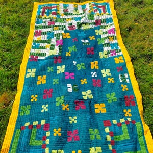 Puede incluir: Una colcha colorida con fondo azul verdoso y borde amarillo. La colcha está compuesta por diversas formas geométricas y patrones, incluyendo cuadrados, rectángulos y flores. La colcha está doblada por la mitad y sobre un lecho de hierba verde.