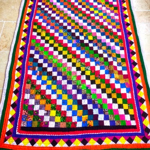 Puede incluir: Una colcha de retazos colorida con un patrón a cuadros en tonos de rojo, azul, verde, amarillo y blanco. La colcha tiene un borde decorativo con formas de diamantes en naranja, amarillo y negro.