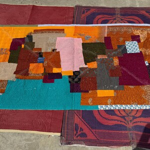 May include: A colourful patchwork quilt featuring orange, teal, pink, and brown squares. The quilt is made from various fabrics sewn together. The rectangular quilt rests on a red and purple patterned rug.