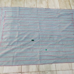 Op de afbeelding: Een rechthoekige, handgemaakte quilt met een blauw-wit geblokt patroon, geaccentueerd door dunne rode strepen. De randen zijn omrand met rode stiksels. Kleine groene en gele patches zijn zichtbaar.
