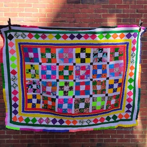 May include: A colourful patchwork quilt with a white background and a chequered pattern. The quilt is made of various fabrics in shades of red, green, blue, yellow, pink, and purple. The quilt is bordered with a diamond pattern in shades of green, yellow, and purple.