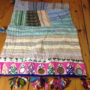 Puede incluir: Una colcha de retazos con un borde rosa y verde, con pequeños espejos y borlas. La colcha está hecha de varias telas en tonos azules, verdes y blancos.