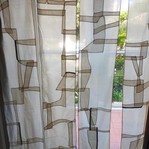 Puede incluir: Cortinas blancas y transparentes con un patrón geométrico de cinta marrón y negra. Las cortinas están colgadas en una ventana con vista a un follaje verde.