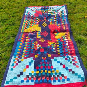 Puede incluir: Un edredón rectangular colorido con un diseño de patchwork. El edredón presenta un panel central azul oscuro con detalles en rojo, amarillo y azul. Los bordes están ribeteados en rojo y azul, sobre hierba verde.