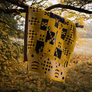 May include: A yellow and black patterned quilt hanging from a tree branch in a forest setting. The quilt has a geometric design with squares and rectangles.