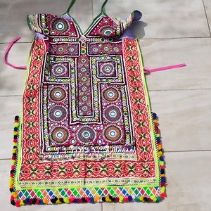 May include: A colourful, embroidered garment with a geometric pattern and mirror embellishments. The garment has a V-neck and a long, flowing skirt. The fabric is a blend of cotton and silk.