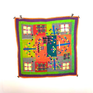 May include: A colorful, square quilt with a patchwork design. The quilt features squares of orange, green, gray, and teal, accented with small squares and lines in various colors. The quilt is bordered with orange, green, and purple. It is hung with metal clips.