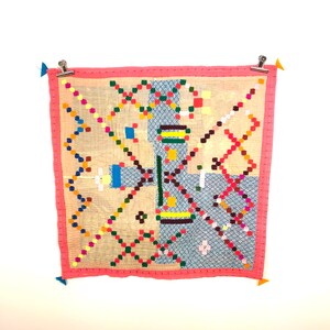 May include: A square, handmade quilt with a light beige background and a pink border. The quilt features a colourful geometric design with squares and lines in various colours, including blue, green, yellow, and red. The quilt is held up by metal clips.