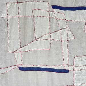 May include: White fabric with red and blue stitching forming geometric shapes. The fabric is textured and has a slightly wrinkled appearance.