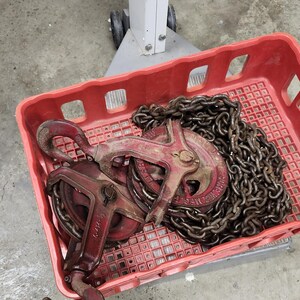 K&ouml;nnte beinhalten: Eine rote Plastikkiste enth&auml;lt zwei antike rote und rostbraune Metallflaschenz&uuml;ge und eine schwere Kette. Die Flaschenz&uuml;ge haben Haken und Herstellerkennzeichnungen. Die Kette ist dick und verbindet die Bl&ouml;cke, was auf einen Hebemechanismus hindeutet.