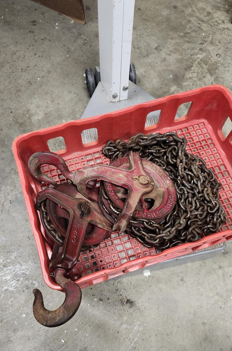 K&ouml;nnte beinhalten: Eine rote Plastikkiste enth&auml;lt zwei antike rote und rostbraune Metallflaschenz&uuml;ge und eine schwere Kette. Die Flaschenz&uuml;ge haben Haken und Herstellerkennzeichnungen. Die Kette ist dick und verbindet die Bl&ouml;cke, was auf einen Hebemechanismus hindeutet.